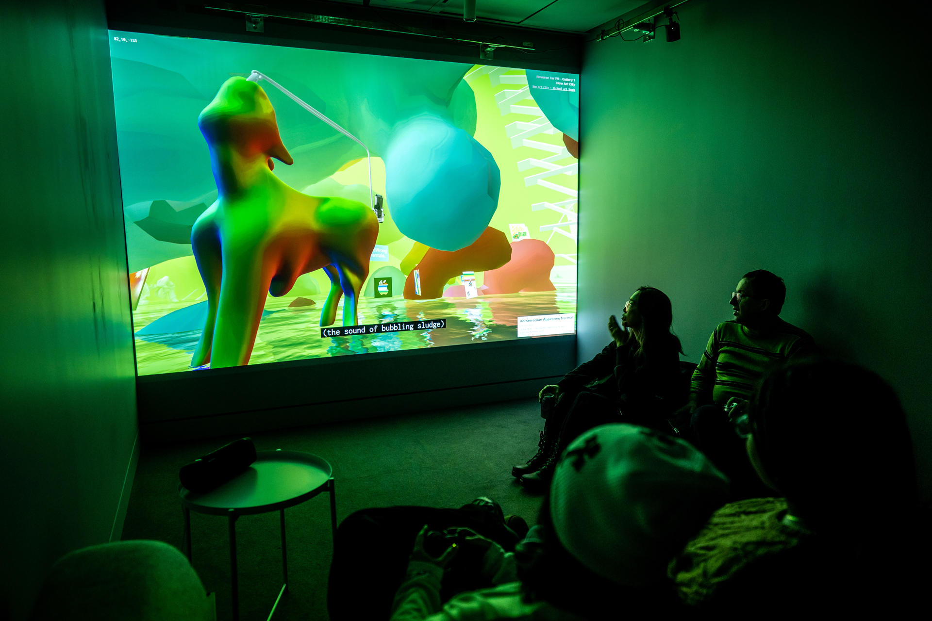 A small room with a couch and beanbag with several visitors viewing the 3D exhibtion on projection and one using a controller'