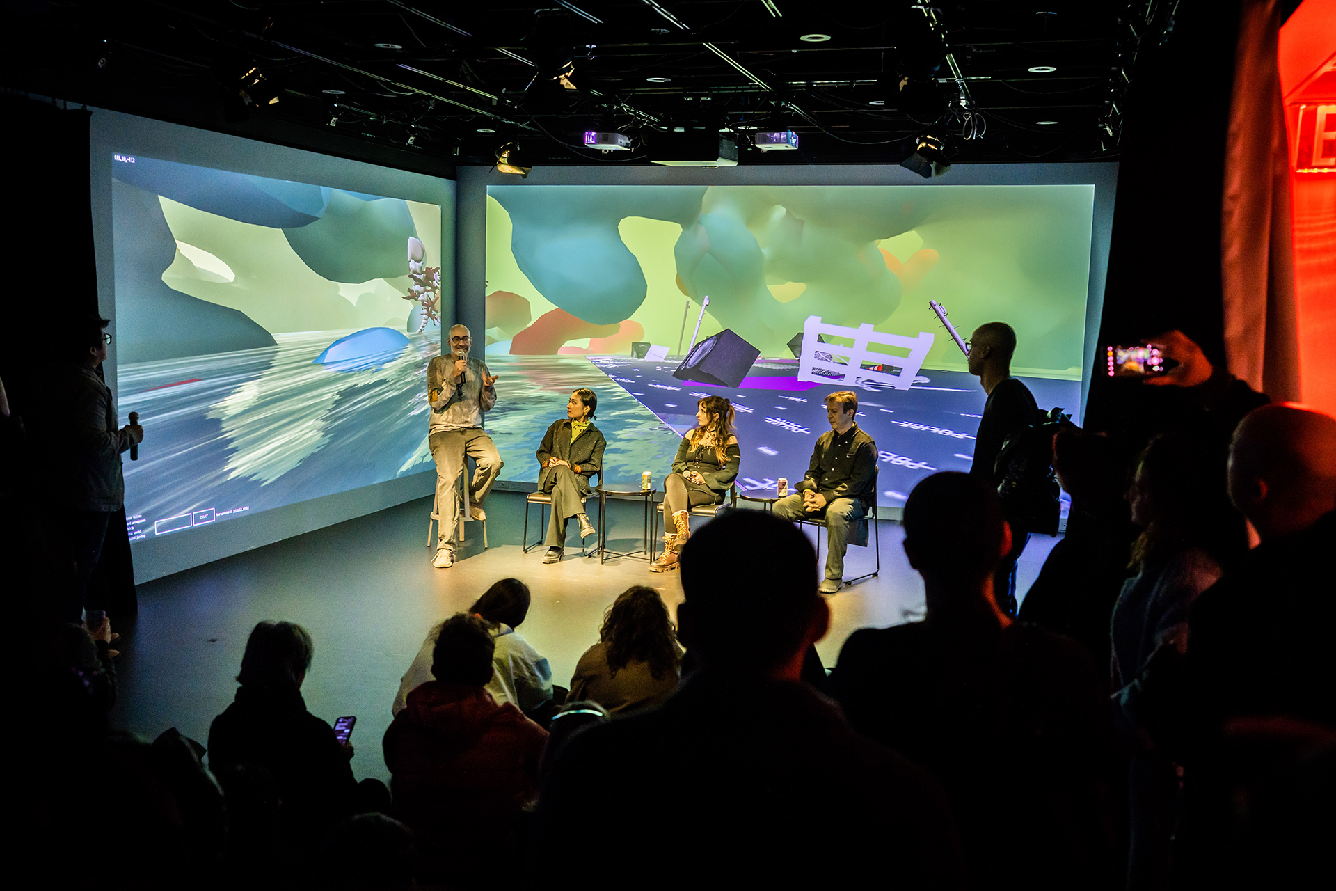 Photograph of four artists seated in a panel discussion style arrangement, with 3d graphics of the exhibition projected behind them.'